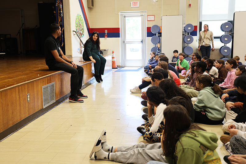 From the side, a young man sits on the edge of a stage while talking to a large number of fifth-grade students.