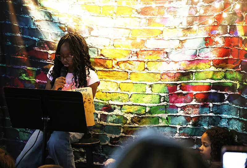 A fourth grade girl reads her poetry. She is in front of a multi-colored brick background.