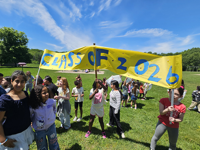 Elementary kids hold up a large blue and gold sign that says Class of 2026