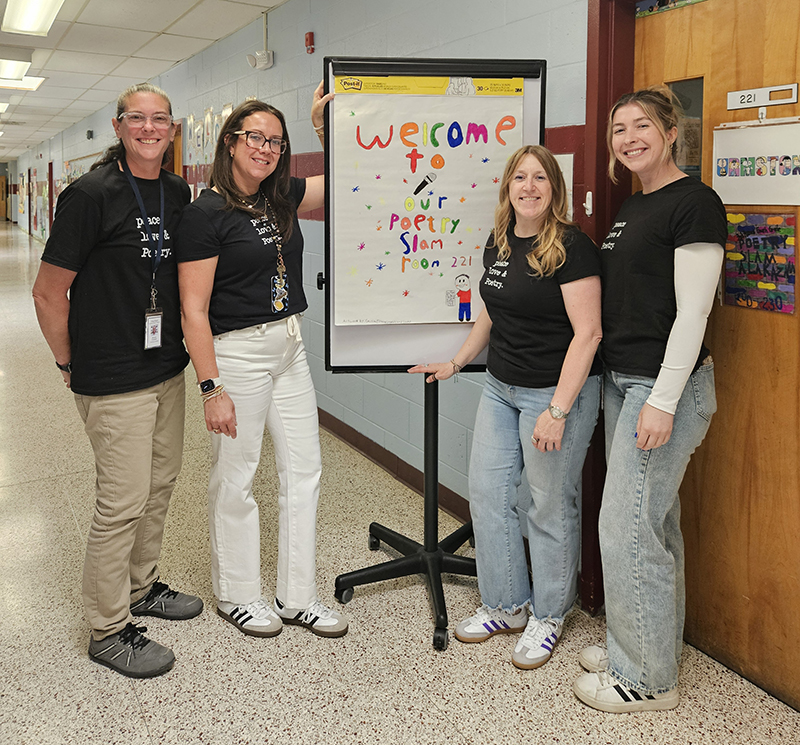 Four women smile. they are all wearing black tshirts that say peace love & Poetry in white. They are standing near a sign that says Welcome to our poetry slam.