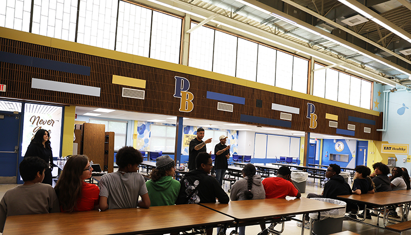 Two young men stand in front of a group of 8th grade students. They are pointing at someone in the crowd.