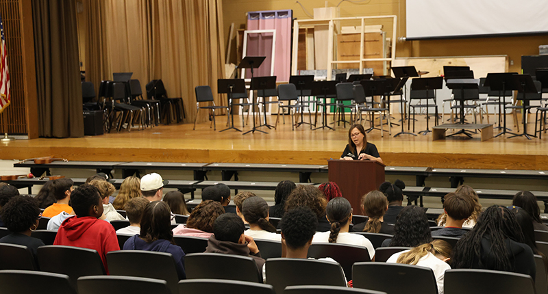 A woman stands in front of a group of 8th grade students. She is in an auditorium at a podium.