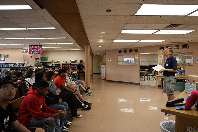A man stands in front of a large group of 8th grade students in a library. He is talking to them about his profession.