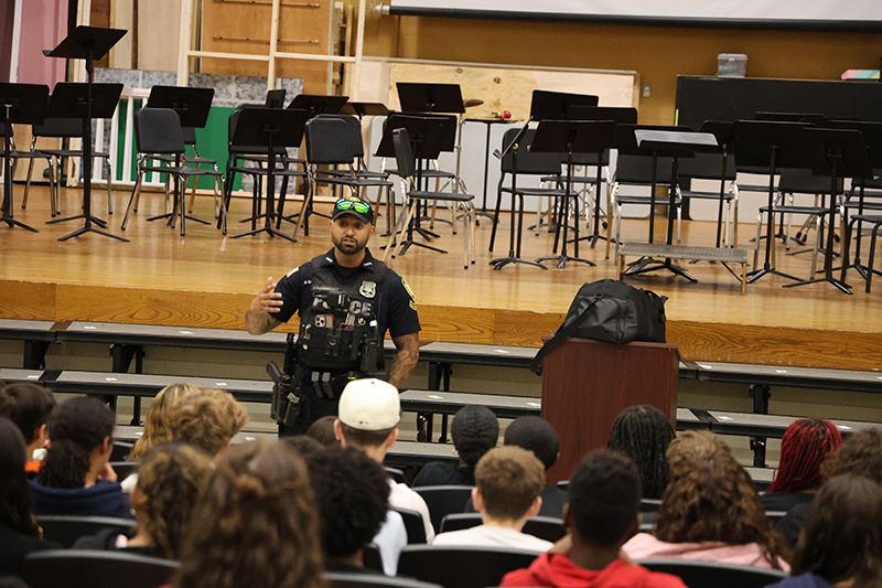 A police officer talks to a group of 8th grade kids in an auditorium.