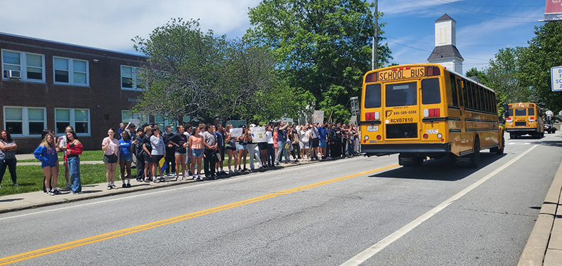 A school bus passes a large group of middle school kids who are cheering.