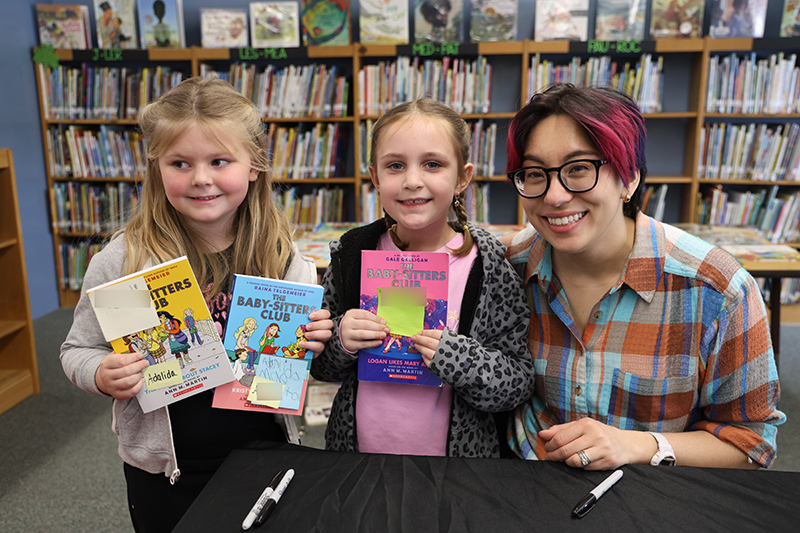 Two smaller elementary students smile and hold books. The person who wrote the books is next to them also smiling.