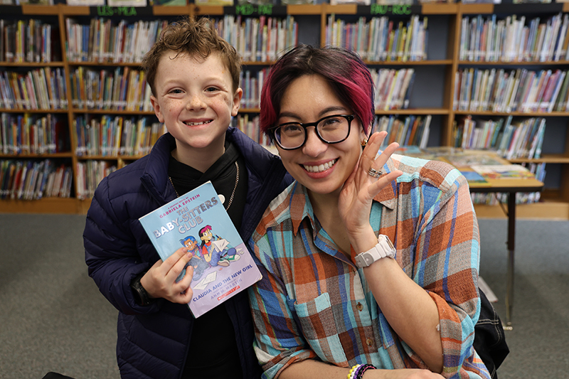 A woman smiles next to a young boy also smiling. He is holding a book that the woman wrote.