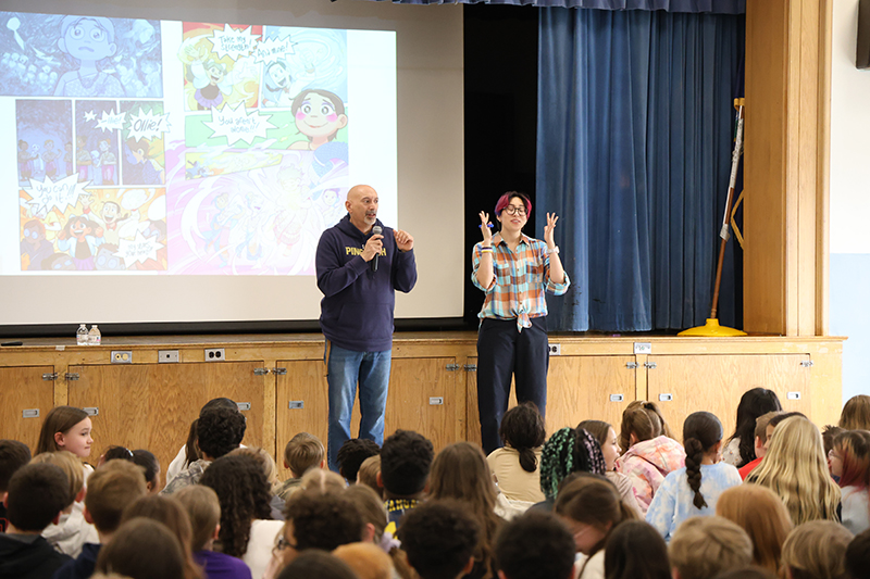 A man and woman stand in front of a large group of elementary kids. In the background are drawings on a screen.