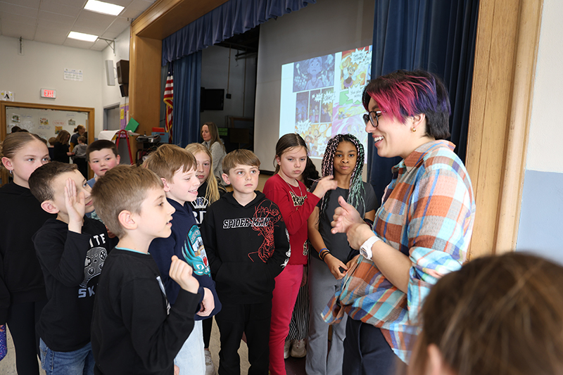 A woman with short dark and red hair talks to a group of elementary kids after giving a talk.