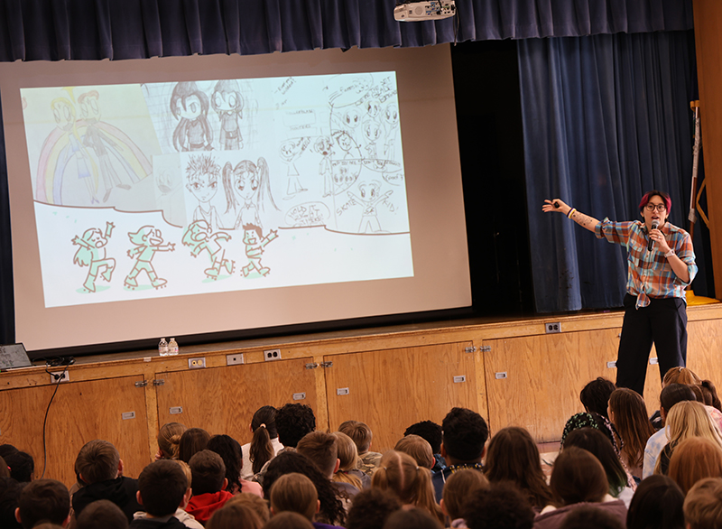 A woman stands in front of a large group of elementary kids and talks to them. She is pointing to a screen that has drawings on it that she produced.