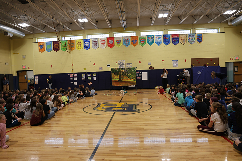 From the back of a large gymnasium, elementary students sit on either side.