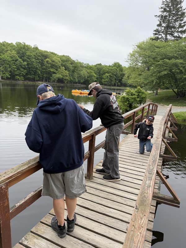A few people - a little kid, high school kid and an adult - fish off a little wooden bridge.