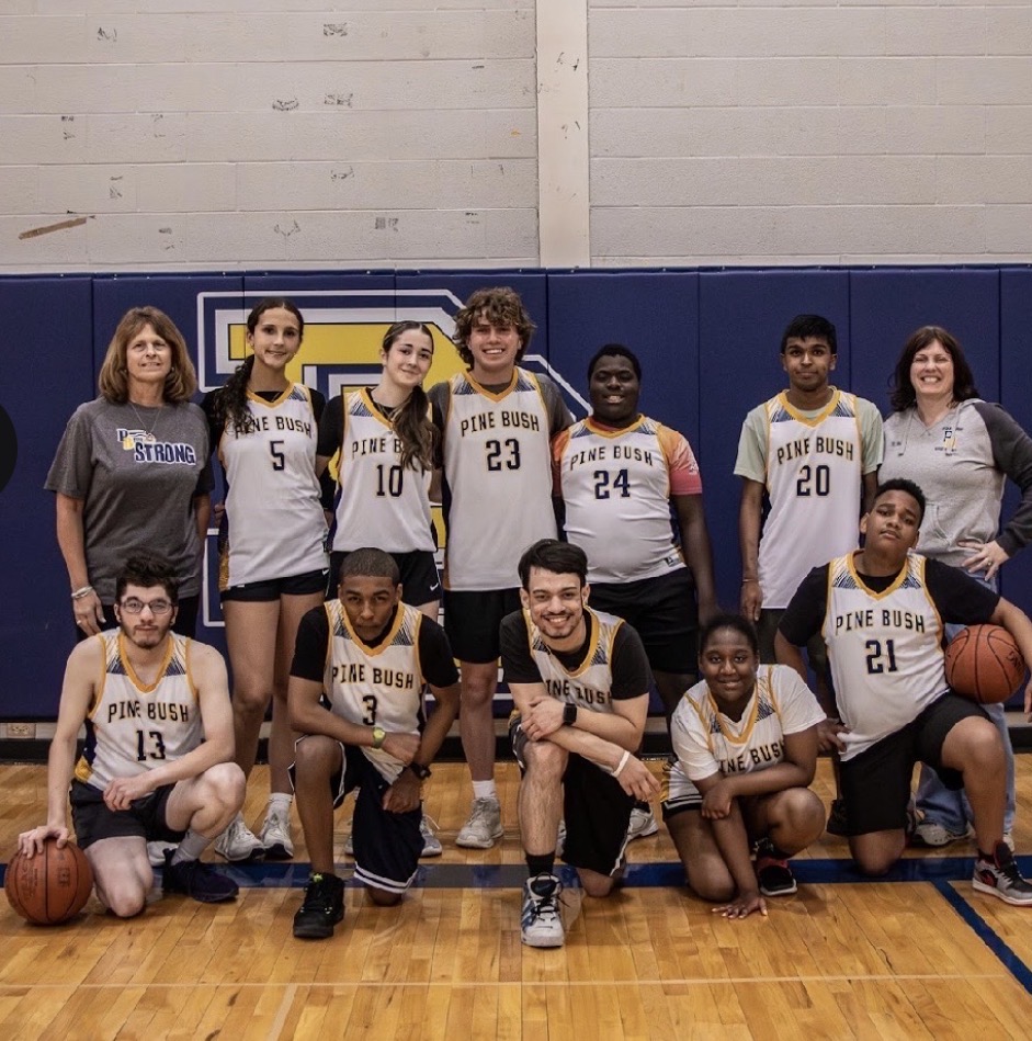 Two rows of basketball players and coaches. The players are wearing white jerseys that say Pine Bush. All are smiling.