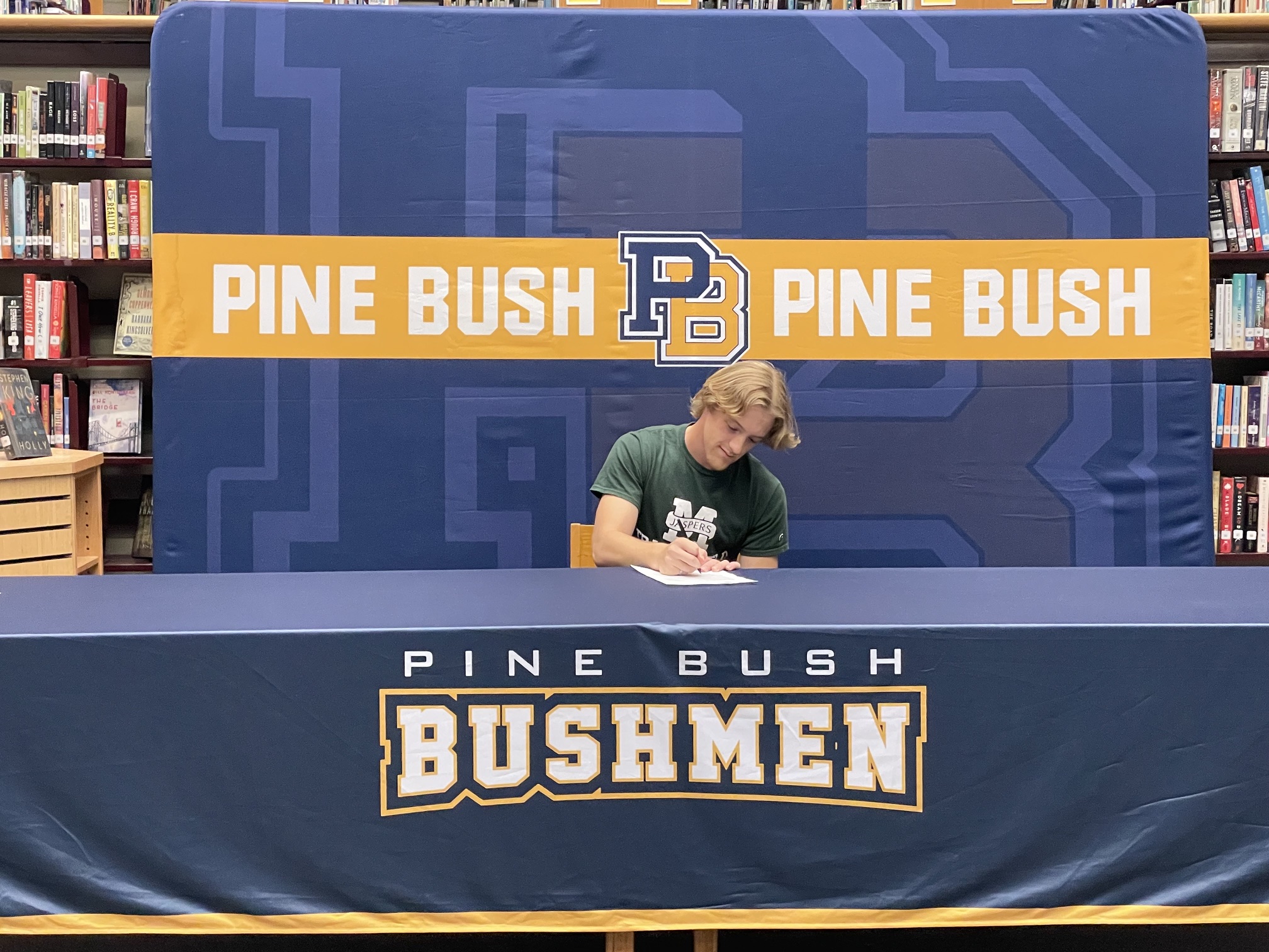A young man wearing a green shirt with Manhattan and a large M across the front sits at a table with Pine Bush Bushmen written across the tablecloth. He signs a piece of paper. 