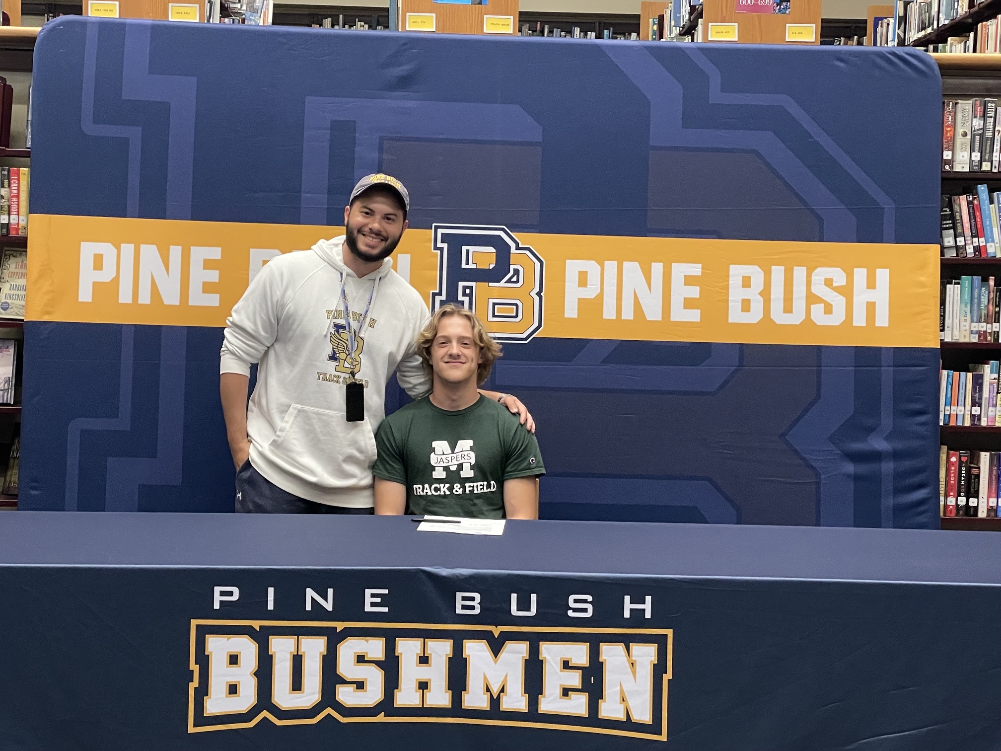A young man wearing a green shirt with Manhattan and a large M across the front sits at a table with Pine Bush Bushmen written across the tablecloth. A man stands with him and they both smile.
