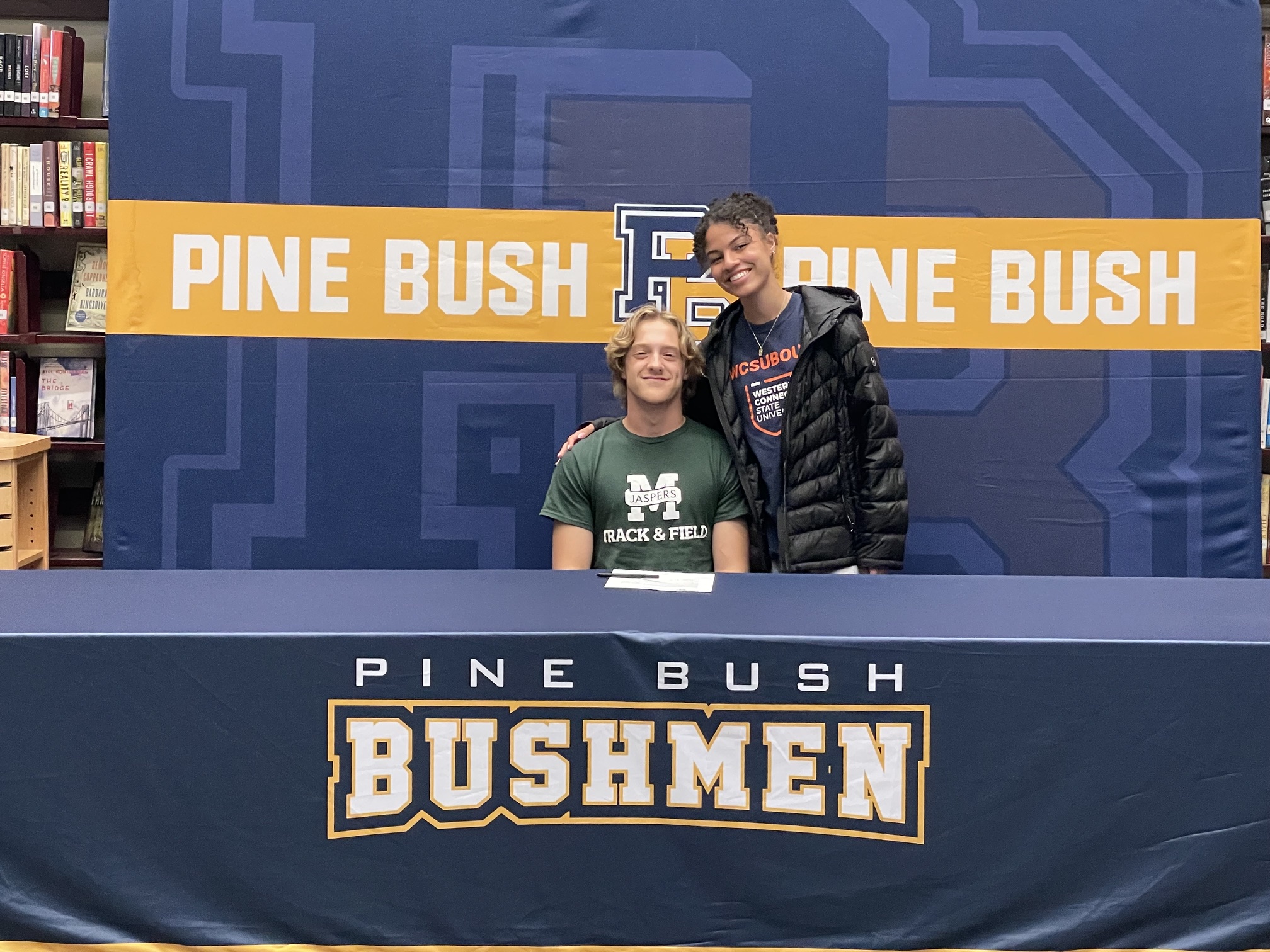 A young man wearing a green shirt with Manhattan and a large M across the front sits at a table with Pine Bush Bushmen written across the tablecloth. A young woman stands with him and they both smile.