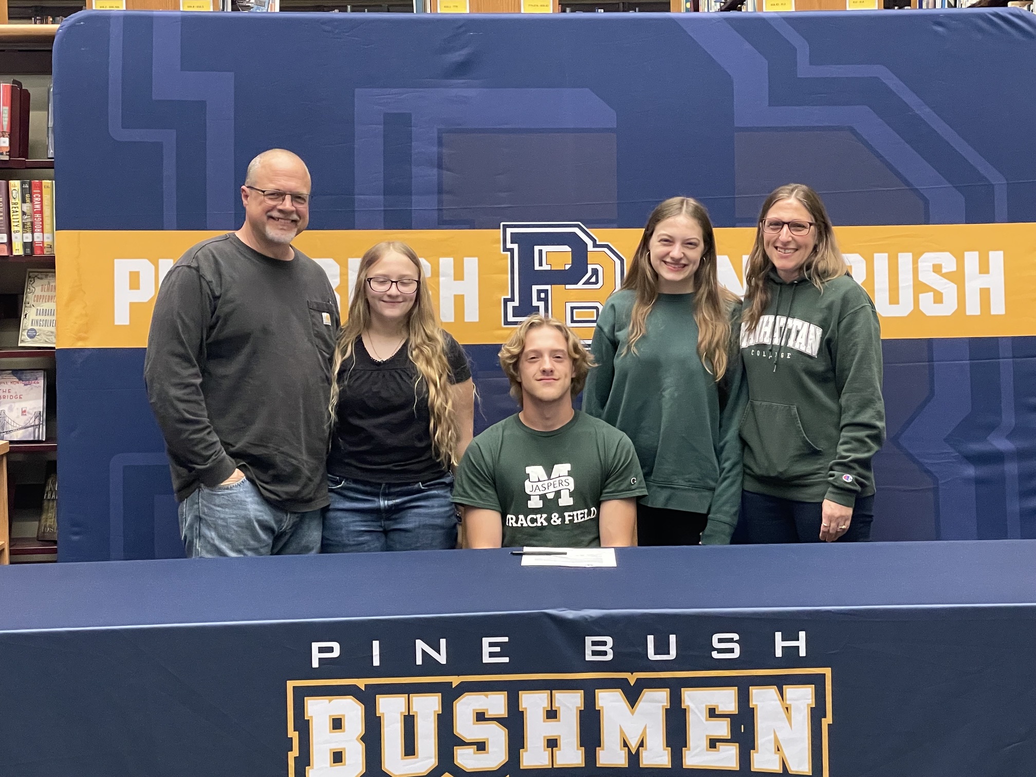 A young man wearing a green shirt with Manhattan and a large M across the front sits at a table with Pine Bush Bushmen written across the tablecloth. Around him are one man, two girls and a woman. All are smiling.
