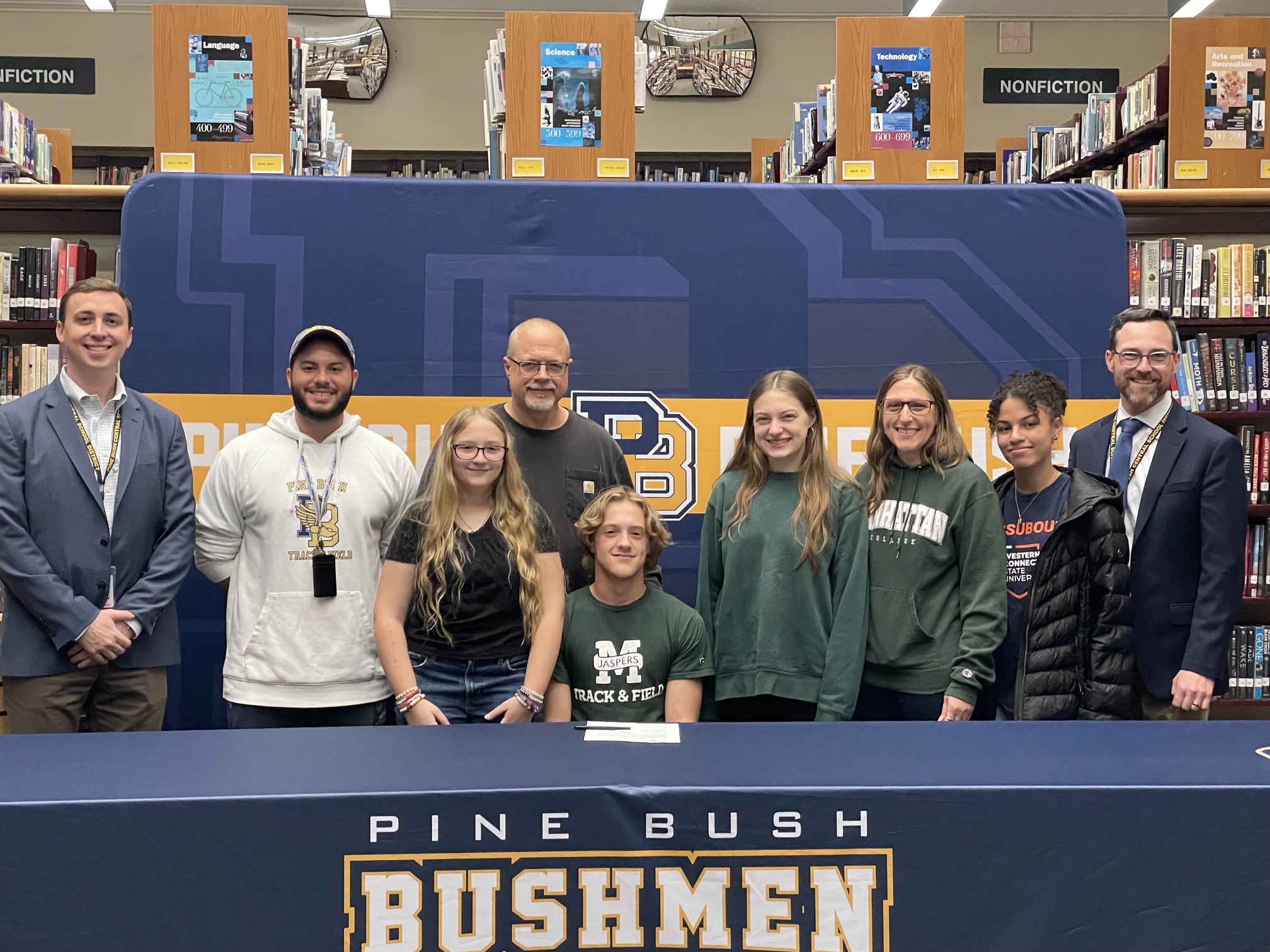 A young man wearing a green shirt with Manhattan and a large M across the front sits at a table with Pine Bush Bushmen written across the tablecloth. Around him are several people. All are smiling.