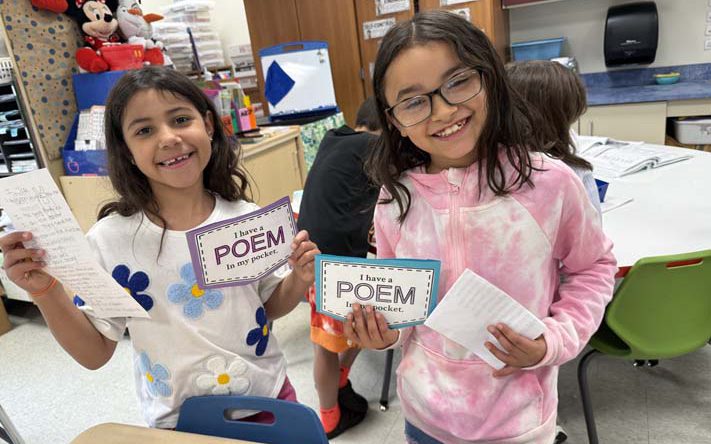 Two elementary age girls smile and hold up small pickets they made that say Poem in Your Pocket.
