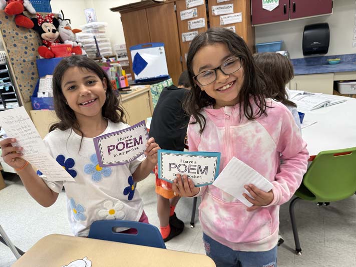 Two elementary age girls smile and hold up small pickets they made that say Poem in Your Pocket.