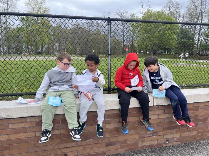 Four elementary age boys sit on a wall . Two read their poems to the boys they are sitting with.