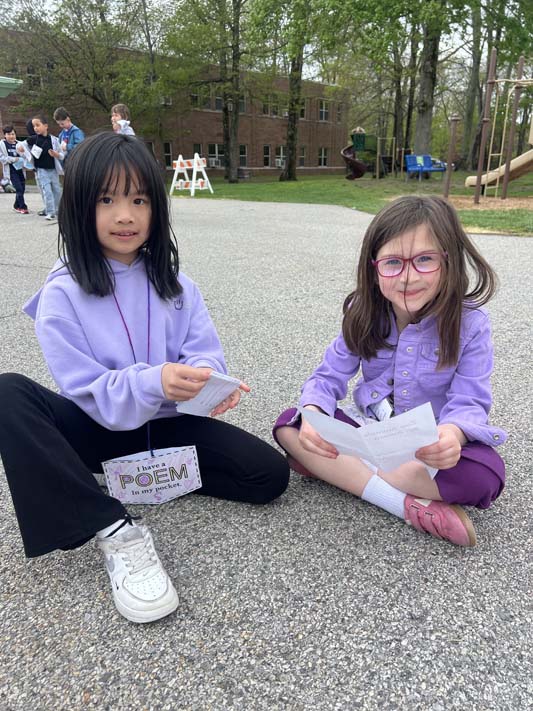 Two elementary age girls sit on the ground and share a poem with  each other.