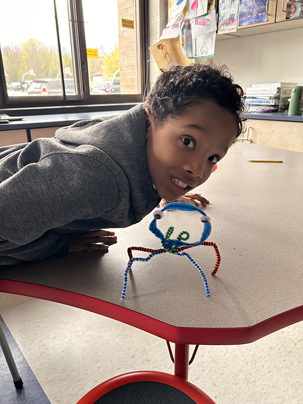 A second-grade boy smiles as he leans in over his pet made from pipe cleaners and googly eyes.