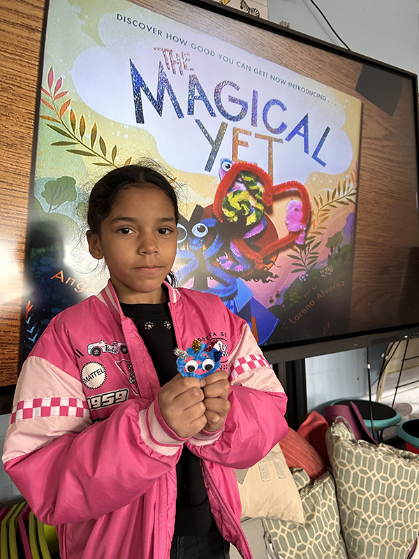 In the background there is a projection of a book called The Magical Yet. In front is a second-grade student holding a pet made from pipe cleaners and googly eyes.