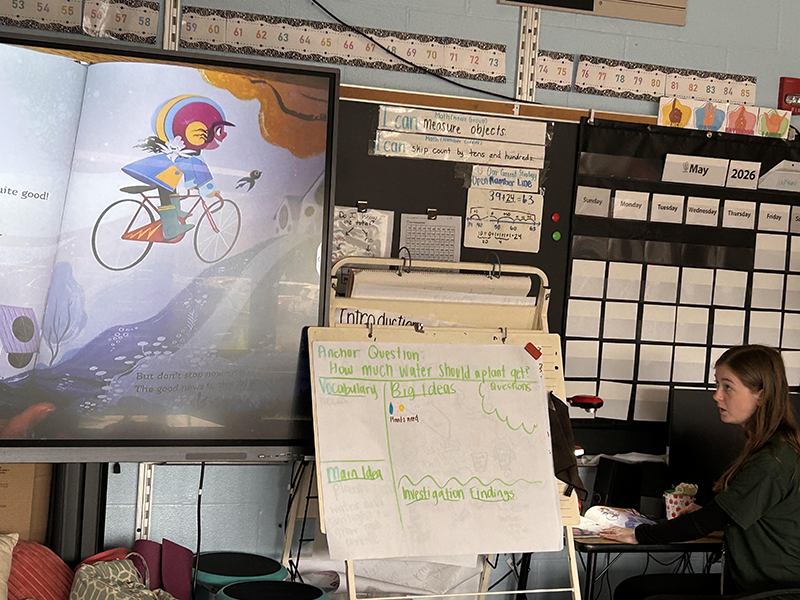 A woman sits by a very large chart. next to her is a projection of a book that is being read to the group.