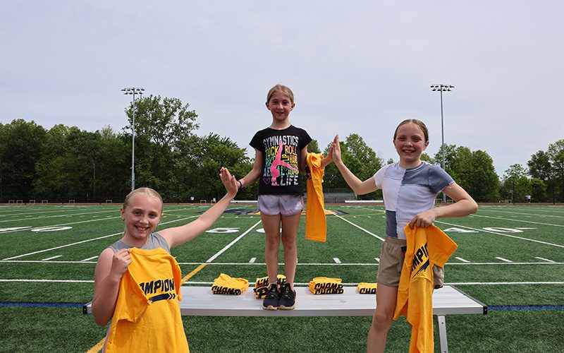 Three elementary girls stand smiling, each holding a yellow tshirt that says champion. They are high fiving.