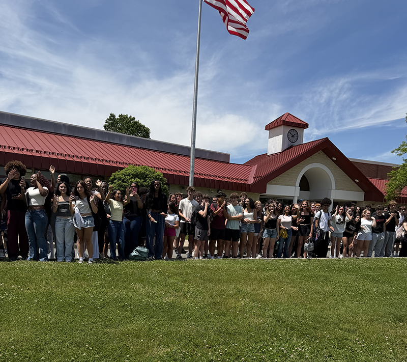 A group of high school kids stand outside of their school cheering.