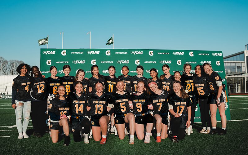 22 high school girls stand in two rows. They are all wearing their blue and gold flag football uniforms. The backdrop is green wit Gatorade ad Jets Flag on it. The sky is beautiful blue.