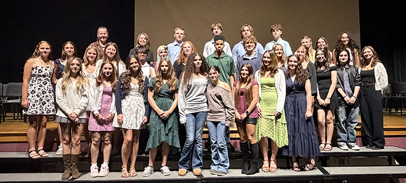 A group of  27 high school students standing in three rows on risers on a stage. Two women also stand with them. all are smiling.