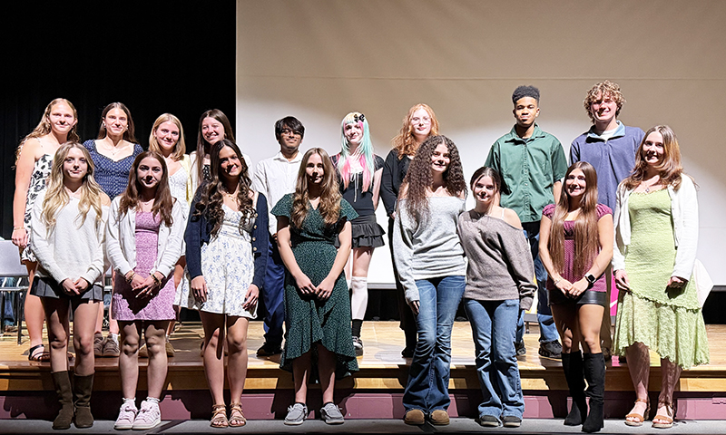 A group of 17 high school student stand in two rows on a stage. They are dressed ni cely and smiling.