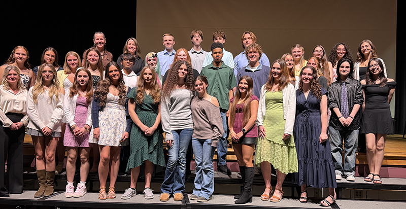 A group of 26 high school students who are smiling stand on a stage. With them are three women as well.
