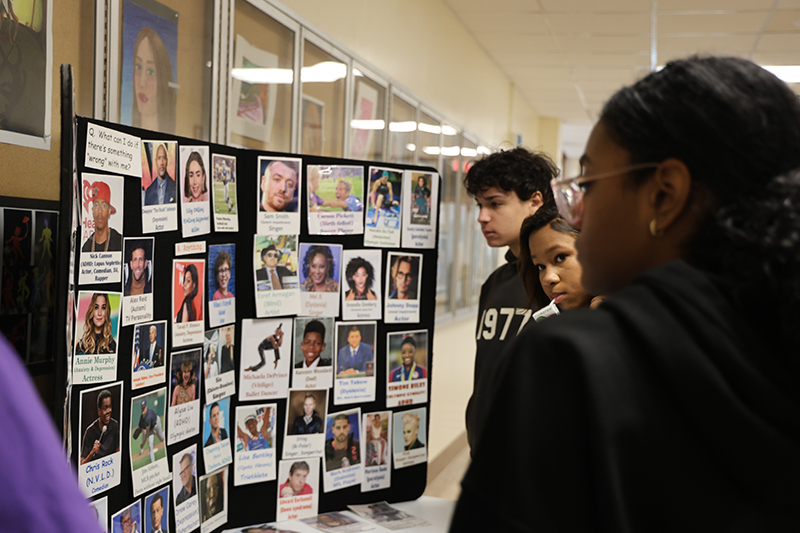 A group of high school kids stand around and look at a large display of photos of famous people who have disabilities.