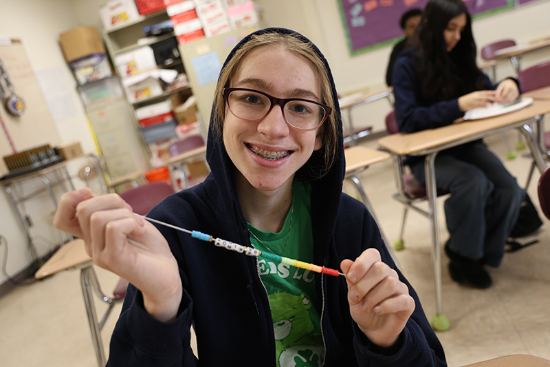 A high school student smiles and holds up a bracelet they just made .