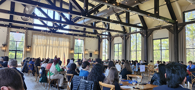 A very large group of people sitting at tables in a nice banquet hall. There is a speaker at the front of the room.