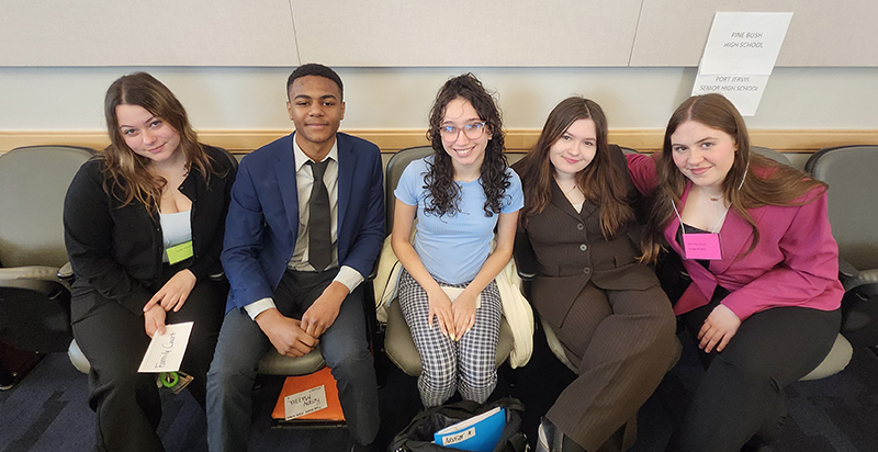 A group of five high school students, dressed nicely, sit on a bench in a building.