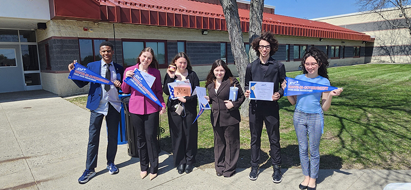 Six high school students stand outside of a large building. Each holds a certificate or pennant.