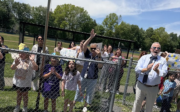 A group of elementary kids wave and a man claps as a bus parade goes by.