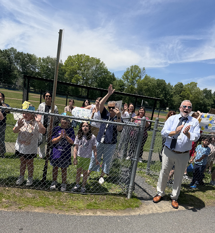 A group of elementary kids wave and a man claps as a bus parade goes by.