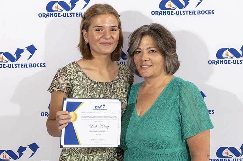 A young woman holds a certificate and smiles. She is standing with a woman in a green dress who is also smiling.