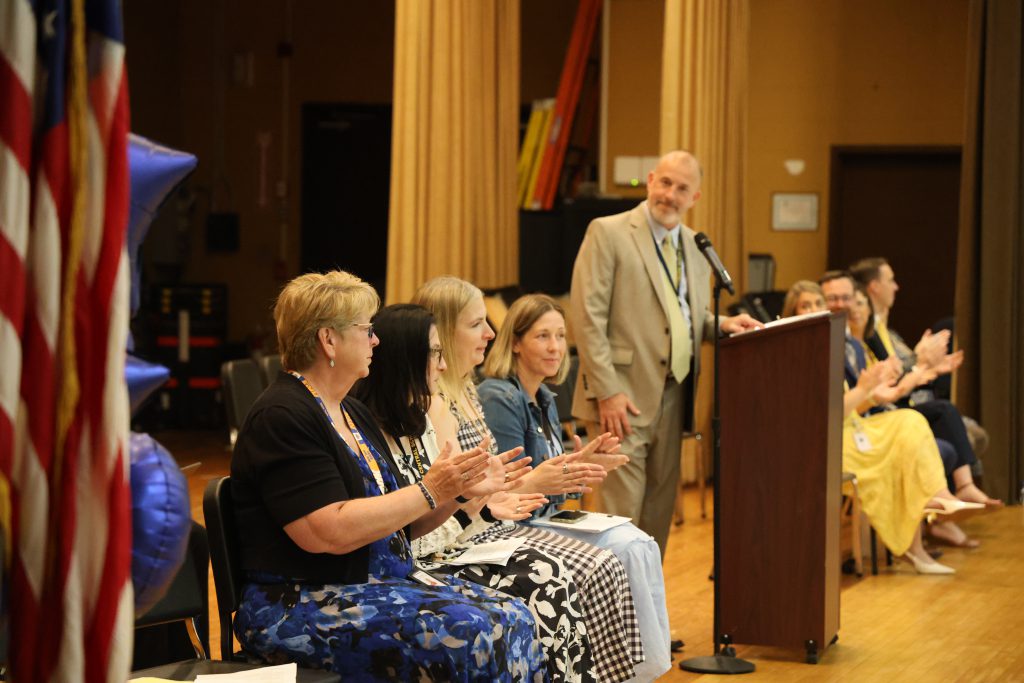 Adults sit on a stage and clap as a man speaks at a podium.