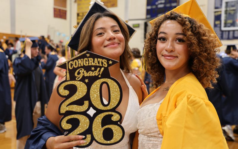 Two high school girls in blue and gold caps and gowns smile. One holds a prop that says congrats grad 2026