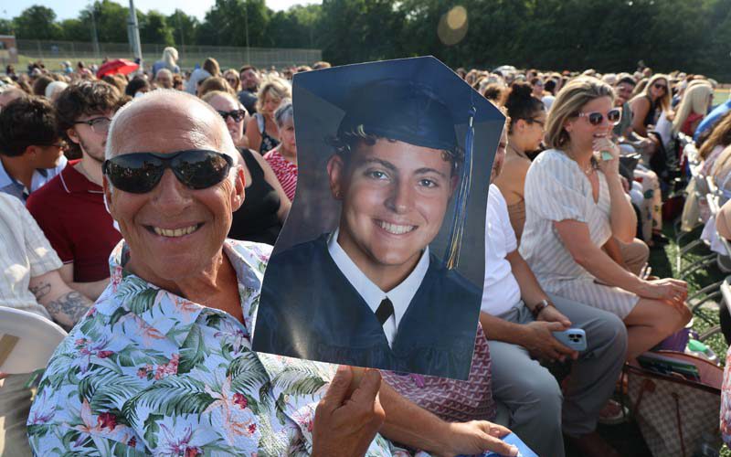 A man smiles as he holds up a large picture of a young man in a cap and gown.