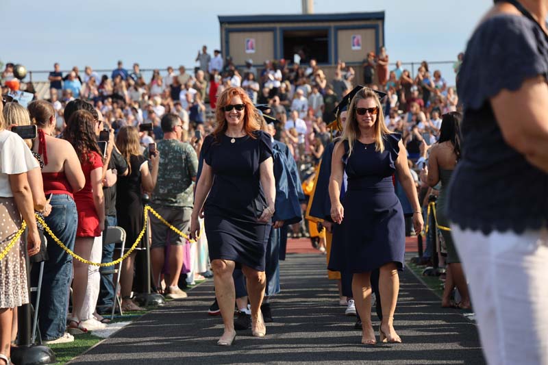 A large group of graduates file onto a football field. Leading the way are two women in blue dresses.