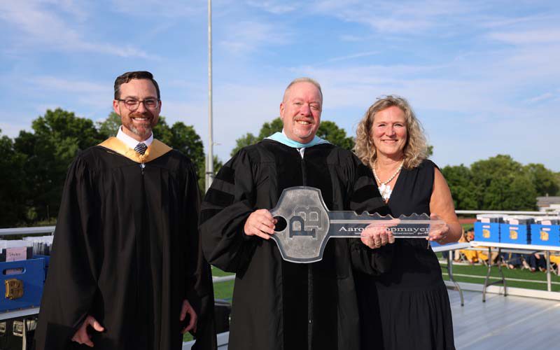 A woman on the right, a man on the left. The man in the center holds a large key that has a PB on top.