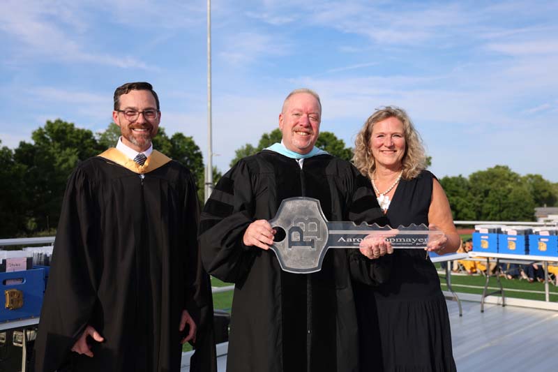 A woman on the right, a man on the left. The man in the center holds a large key that has a PB on top.
