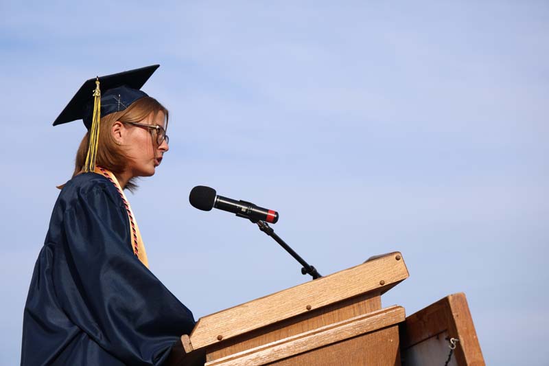 A young woman with short blonde hair and glasses wearing a navy blue cap and gown speaks at a podium.